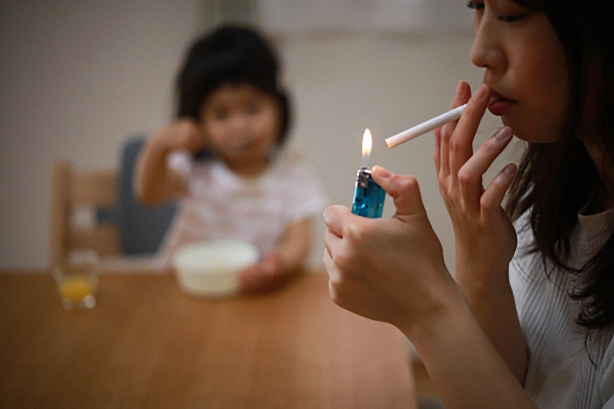 Image of a lady holding a cigarette and lighter who is about to smoke sitting with her daughter in background who is blurred in the picture.